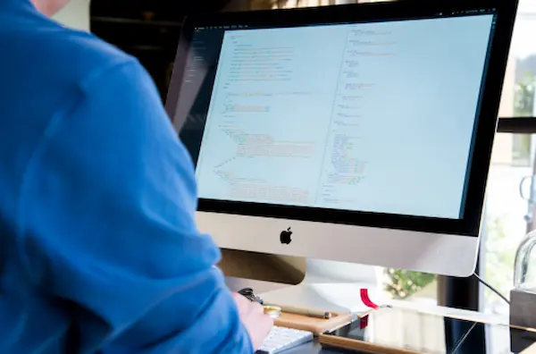 Eine Person von hinten, die vor einem großen Apple iMac-Bildschirm sitzt, auf dem Codezeilen angezeigt werden. Die Person scheint zu arbeiten, mit ihrer Aufmerksamkeit auf den Bildschirm gerichtet. Die Umgebung deutet auf ein Zuhause oder ein zwangloses Büro hin, mit natürlichem Licht, das von einem Fenster kommt. Die Person trägt ein blaues Hemd, und auf dem Schreibtisch liegen ein Stift und ein Notizbuch, was auf eine Arbeits- oder Lernumgebung hindeutet. Der Fokus auf den Bildschirm mit Code lässt vermuten, dass die Person wahrscheinlich in der Programmierung oder Softwareentwicklung tätig ist