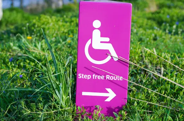 Ein magentafarbenes Schild mit einem weißen Piktogramm einer Person im Rollstuhl und einem Pfeil nach rechts, mit dem Text 'Stufenfreier Weg' vor einem Hintergrund aus grünem Gras.